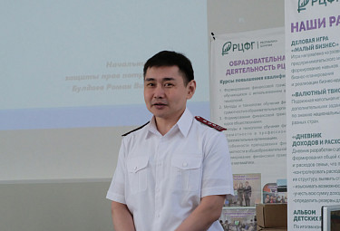Роман Булдаев на фоне экрана со своим именем и стендов с материалами о финансовой грамотности (2023 год)